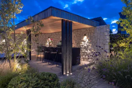 Overdekte buitenruimte met natuurstenen wand, warme buitenverlichting en sfeervolle tuinbeplanting in de avond.