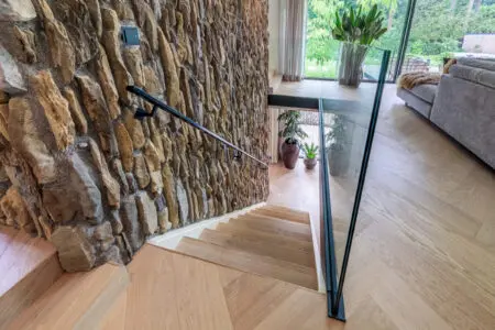 Detail van verticale natuursteen wand binnenshuis, toegepast langs een trap met glazen balustrade.