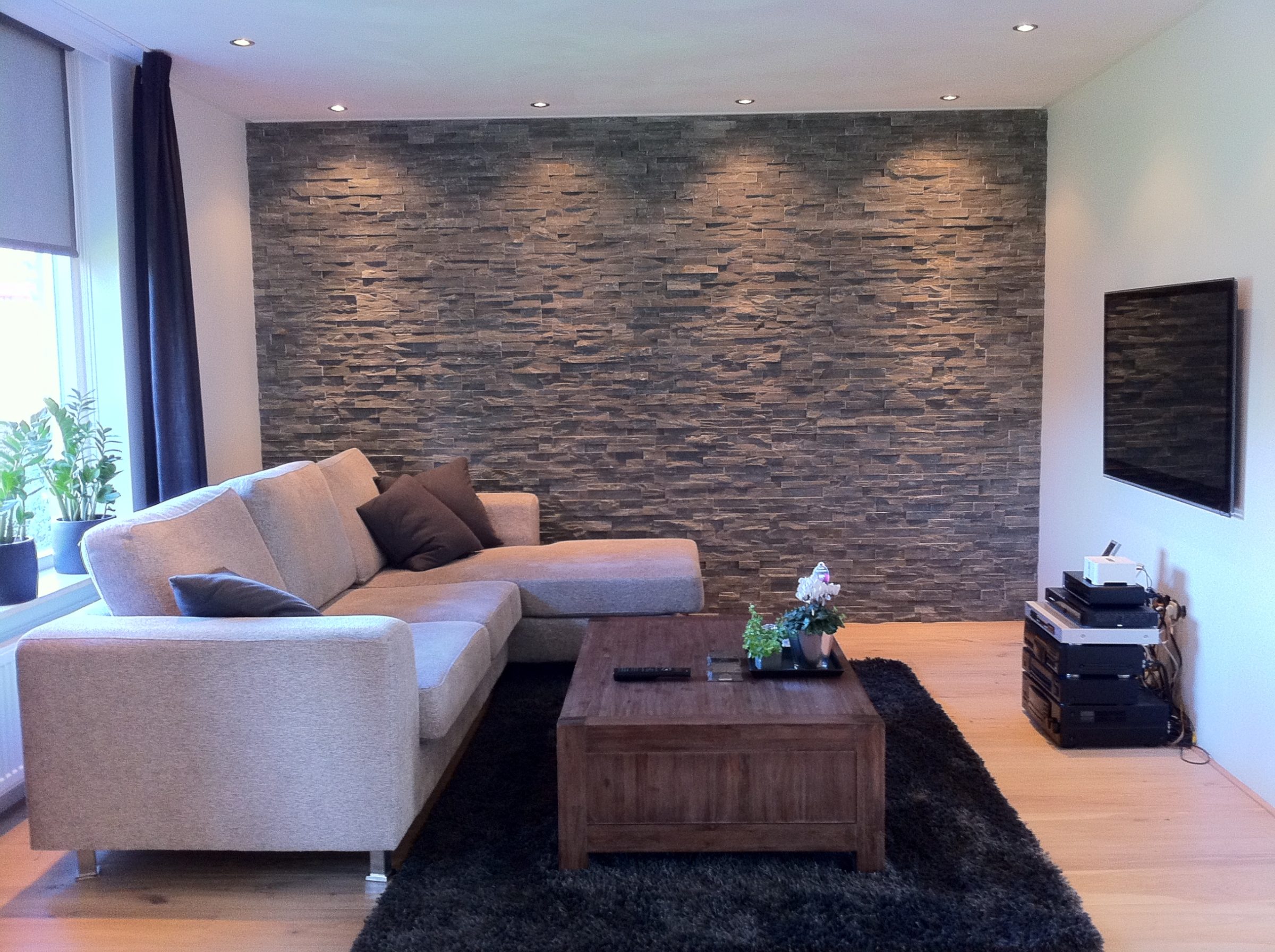 Sfeervolle woonkamer. De wand is bekleed met Grijs Kwartsiet Natuursteenstrips. Deze strips zijn zowel voor binnengebruik als buitengebruik geschikt.